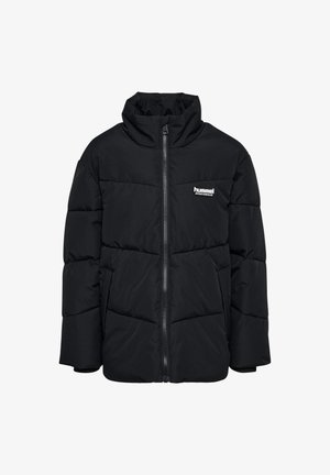 Enfant portant un bonnet en laine marron, une doudoune noire, un pantalon de survêtement gris et des baskets blanches, marchant sur un fond blanc uni.