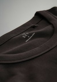 Camiseta de algodón marrón oscuro con textura acanalada, con cuello redondo y una etiqueta que dice "THE SET M HECHO EN BANGLADESH" dentro del cuello.