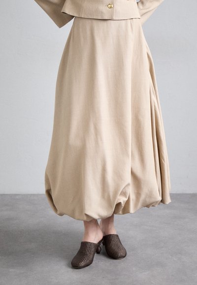 Gonna beige fluida con un ampio design plissè, abbinata a sabot marroni texturizzati. Il tessuto appare morbido e leggero.