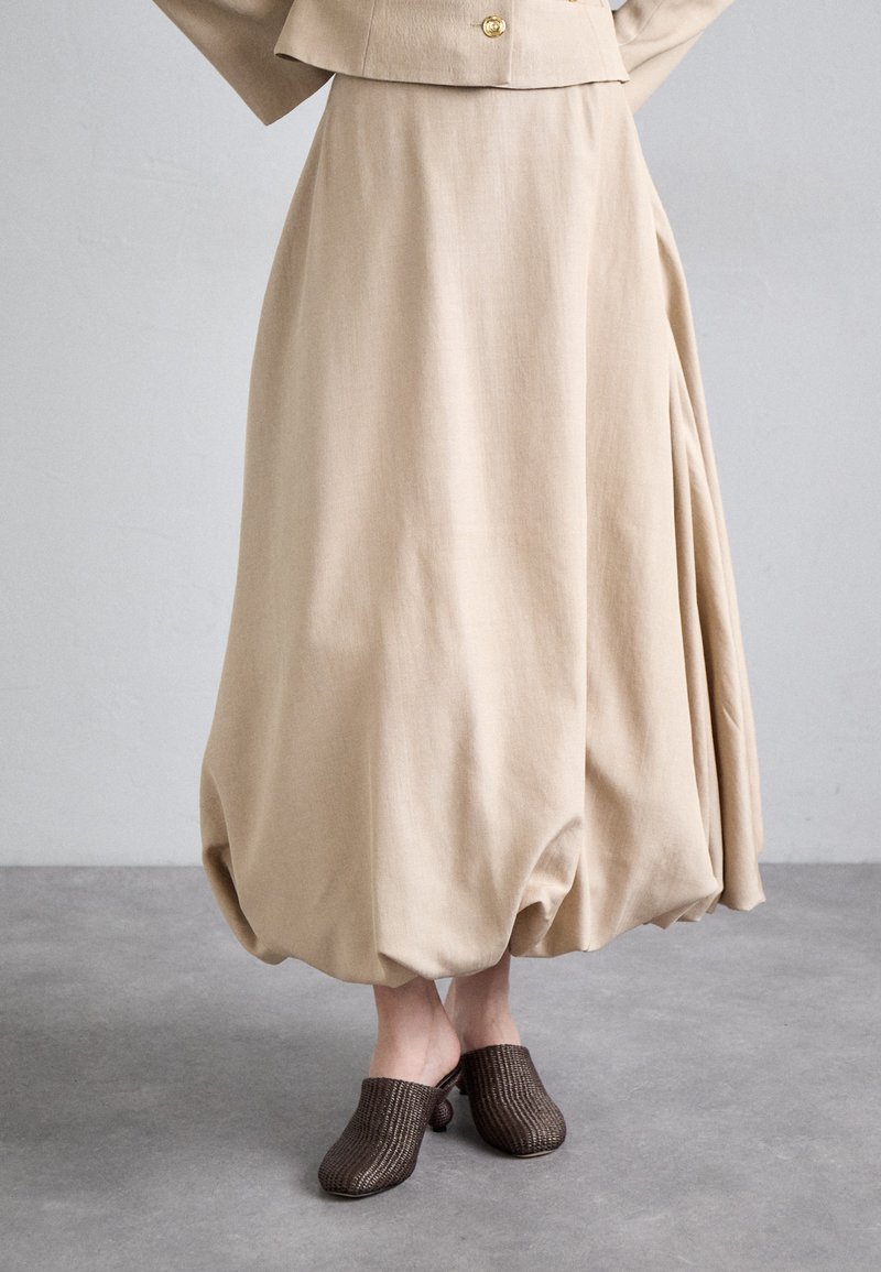 Jupe beige fluide avec un large design plissé, associée à des sabots en cuir texturé marron. Le tissu semble doux et léger.