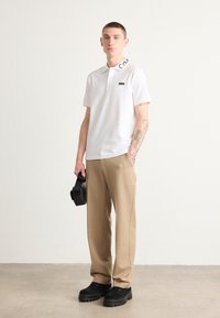 Witte polo met korte mouwen en zwart logo detail op de kraag, gecombineerd met beige broek en zwarte chunky schoenen. Houdt een zwarte tas vast.