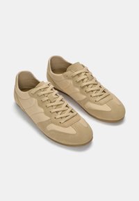 Baskets de couleur beige en cuir lisse avec un bout arrondi, des lacets plats et de légères textures sur les côtés.