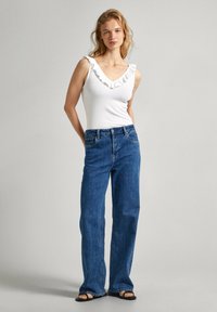 Top bianco con scollo arricciato abbinato a jeans dritti in denim blu. La modella sta in piedi con le mani nelle tasche, indossando sandali neri.