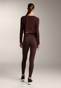 Nainen, jolla on poninhäntä, pukeutuneena matching-tumma ruskea pitkähihainen cropped-paita, jossa on selkäläppä, ja korkea vyötärö leggingit, seisoo yksinkertaista taustaa vasten.