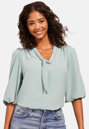 Blusa azzurra chiara con colletto a nodo, maniche corte a palloncino e texture morbida. Abbinata a pantaloni corti in denim, con una vestibilità rilassata.
