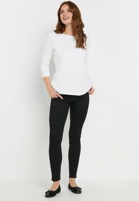 Weißes langärmliges Oberteil mit einem Bootsausschnitt, kombiniert mit schwarzen Skinny-Jeans und glänzenden schwarzen Ballerinas mit einer Schleifenverzierung.