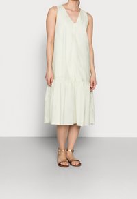 Hellgrünes ärmelloses Kleid mit V-Ausschnitt, gerafftem Bund und weitem Saum. Model kombiniert es mit hellbraunen Riemchensandalen. Weicher, fließender Stoff.