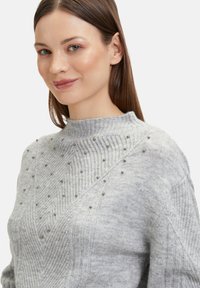 Helles graues Sweatshirt mit geripptem Material, versehen mit dekorativen silbernen Nieten, die in einem Muster auf der oberen Vorderseite und den Schultern angeordnet sind.