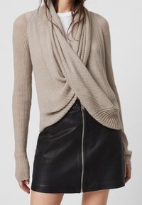 Suéter envolvente de punto beige con mangas largas, que presenta un frente drapeado y se combina con una falda de cuero negra con cremallera. Textura suave y diseño ajustado.