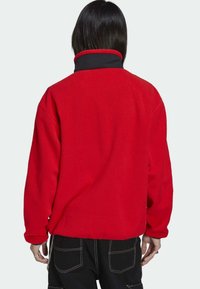 Persona con cabello negro vistiendo una chaqueta de forro polar roja con cuello negro, pantalones negros con costuras blancas, de pie de espaldas.