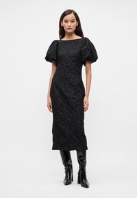 Črna obleka s puhadnimi rokavi, teksturiran cvetlični vzorec, prilegajoča silhueta, do kolen, v kombinaciji s črnimi škornji do kolen. Preprost izrez.