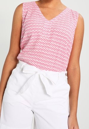 Ärmelloses, rosa gemustertes Oberteil kombiniert mit hoch taillierten weißen Shorts, die am Bund mit einem Stoffgürtel gebunden sind.