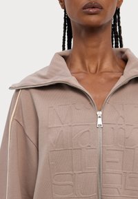 Giacca marrone con zip e colletto alto, caratterizzata da testo impresso e piping beige a contrasto lungo le maniche. Tessuto morbido e textured.