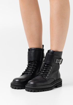 Bottines en cuir noir avec semelles épaisses, lacets sur le devant, large bride avec boucle argentée, portées sur des jambes nues sur fond blanc.