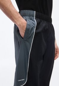 Άνδρας που φοράει αθλητικές παντελόνες Calvin Klein σε μαύρο και σκούρο γκρι με ελαστική μέση, το χέρι του ακουμπάει στην πλαϊνή τσέπη.