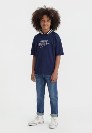 Kind met krullend haar dat een navy Nike polo draagt, opgerolde blauwe jeans en witte hoge sneakers, staand en glimlachend.
