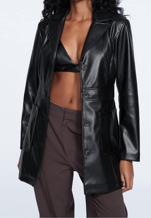 Vrouw die een lange blazer van zwart nepleer draagt over een zwarte bralette en losse bruine broek, gefotografeerd tegen een effen achtergrond.