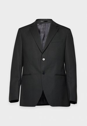 Un homme se tient devant un fond uni, vêtu d’un costume noir, d’une chemise blanche, d’une cravate à pois et de chaussures noires.