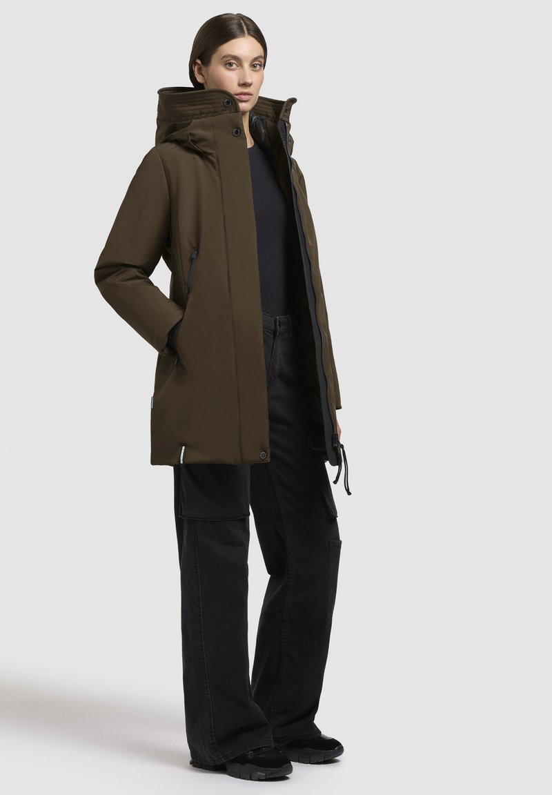 Parka aislante de color verde oscuro con un alto cuello, cierre de cremallera, bolsillos laterales y cordón ajustable, combinada con pantalones cargo negros y zapatos.