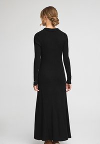 Schwarzes, geripptes Maxikleid mit langen Ärmeln und taillierter Passform. Besitzt einen dezenten Kragen und Schlitte an den Bündchen und besteht aus einem strukturierten Stoff.