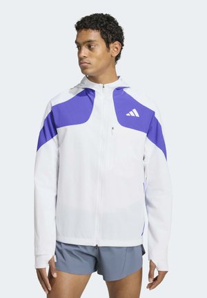 Giovane uomo che indossa una giacca con zip bianca e viola con cappuccio e pantaloncini sportivi grigi, guarda di lato su uno sfondo semplice.