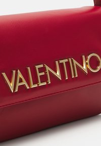 Červená kožená kabelka s výrazným zlatým kovovým logem "VALENTINO", hladkou strukturou a propracovaným designem s prošívanými detaily.