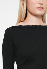 Top negro de canalé con mangas largas que presenta un escote de encaje en forma de ondas, textura suave y silueta ajustada.