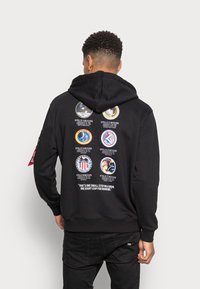 Sweatshirt preto com cordão, apresentando vários emblemas coloridos da missão Apollo nas costas e uma citação na parte inferior. Tecido de textura suave.