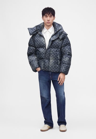 Jeune homme portant une doudoune sombre à motifs par-dessus une chemise blanche avec un jean bleu et des baskets blanches, debout devant un fond uni.