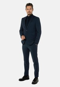 Donkerblauw pak van gladde stof, nauwsluitende pasvorm met een notch revers, gecombineerd met een zwart shirt en glanzende zwarte instappers.