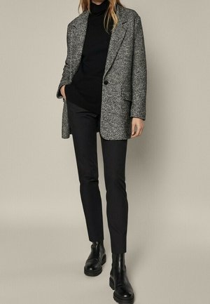 Vrouw die een grijs gestructureerd colbert draagt, een zwarte coltrui, zwarte smalle broek en zwarte chunky leren enkellaarzen, staand met één hand in de zak.