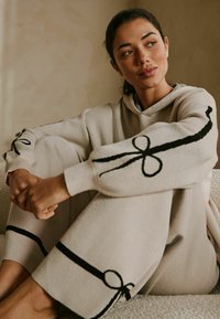 Beige loungewear sæt lavet af blødt stof, med sorte broderede detaljer og sløjfedesign på ærmerne og manchetterne. Løs pasform, hætte trøje.