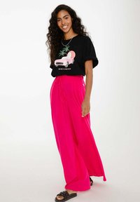 Usmívající se žena s dlouhými kudrnatými vlasy má na sobě černé tričko s potiskem růžového auta a jasně růžové široké kalhoty, stojí s jednou nohou vpřed.