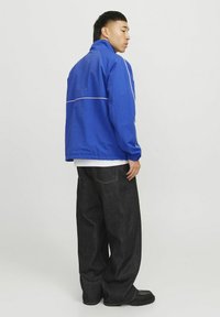 Blå vindjakke med hvide detaljer, parret med mørke, løstsiddende jeans og sort fodtøj, set fra en side- og bagvinkel.