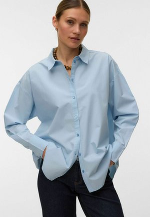 Femme portant une chemise oversize bleu clair avec poches, pantalon foncé et un collier de perles sur un fond blanc uni.