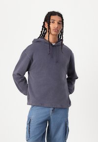 Hanorac oversized de culoare violet, din material moale, cu buzunar tip kangur, glugă cu șnur ajustabil și manșete cu nervuri. Asortat cu pantaloni scurți cargo albaștri.