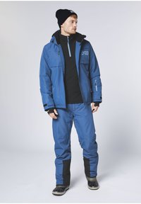 Blaue isolierte Jacke mit schwarzen Akzenten, Reißverschlusstaschen und hohem Kragen, kombiniert mit passenden blauen Hosen und robusten schwarzen Schneestiefeln.