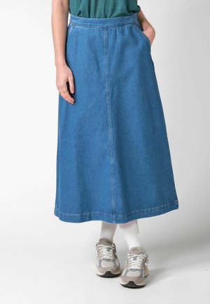 Osoba nosi modrú denimovú sukňu po lýtka, biele ponožky, sivé a biele tenisky a zelený top, pričom má jednu ruku v vrecku.