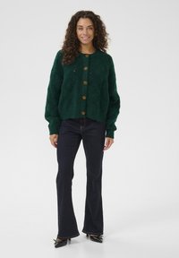 Zaļš adīts kardigāns ar teksturētu rakstu, apaļu kakla izgriezumu un pieciem lieliem zelta pogām. Stilizēts ar tumšiem platām kājām džinsiem un melniem papēžiem.