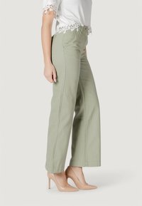 Pantaloni a gamba larga verde chiaro in cotone, con cuciture frontali e chiusura a zip. Abbinati a tacchi alti nudi e a un top bianco con pizzo.