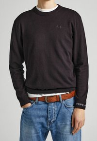 Muž v tmavě hnědé mikině Pepe Jeans přes bílou košili, s hnědým páskem a modrými džíny, se zhandy v kapsách a viditelnými logy značky.