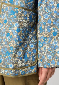 Giacca a fiori con un mix di fiori blu, bianchi e beige su sfondo marrone, con rifiniture verdi lungo i bordi e le tasche.