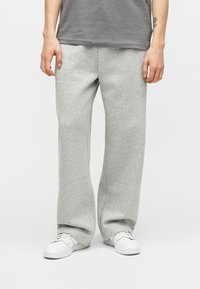 Άτομο που φοράει ανοιχτό γκρι φαρδιά παντελόνια φόρμας, λευκά αθλητικά παπούτσια και γκρι πουκάμισο, στέκεται μπροστά σε απλό φόντο.