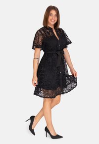 Robe en dentelle noire avec des manches courtes et évasées, un devant à boutons et une ceinture à la taille. Présente des motifs floraux complexes et un ourlet en forme de vagues.