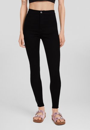 Leggings negros de talle alto hechos de una tela elástica, con cierre de botón delantero, combinados con sandalias planas de color rosa claro.
