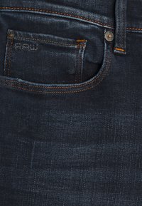 Mörkblå denimjeans med en framficka som har orange sömnad och metallbeslag. Texturen visar liten blekning och en slät yta.