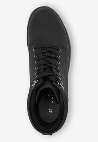 Botín negro con cordones hasta el tobillo, con un acabado de cuero suave, ojales de metal y un cuello acolchado. El interior presenta un suave forro negro.