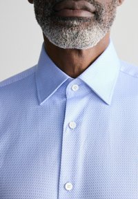 Himmelblaues, knöpfbares Oberhemd mit strukturiertem Rautenmuster, klassischem Kragen und weißen Knöpfen, das das Erscheinungsbild eines glatten Stoffes betont.