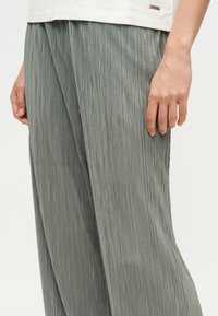 Κοντινό πλάνο ενός ατόμου που φοράει χαλαρό, κάθετα πιετά γκρι παντελόνι και λευκό τοπ με εμφανές χέρι που ακουμπάει στο πλάι.