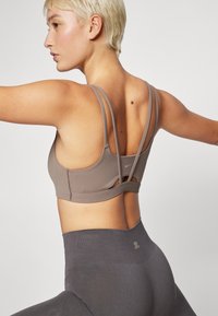 Sujeción deportiva marrón con múltiples tirantes delgados, un acabado texturizado y un detalle recortado en la parte posterior. Leggings gris de cintura alta visibles.